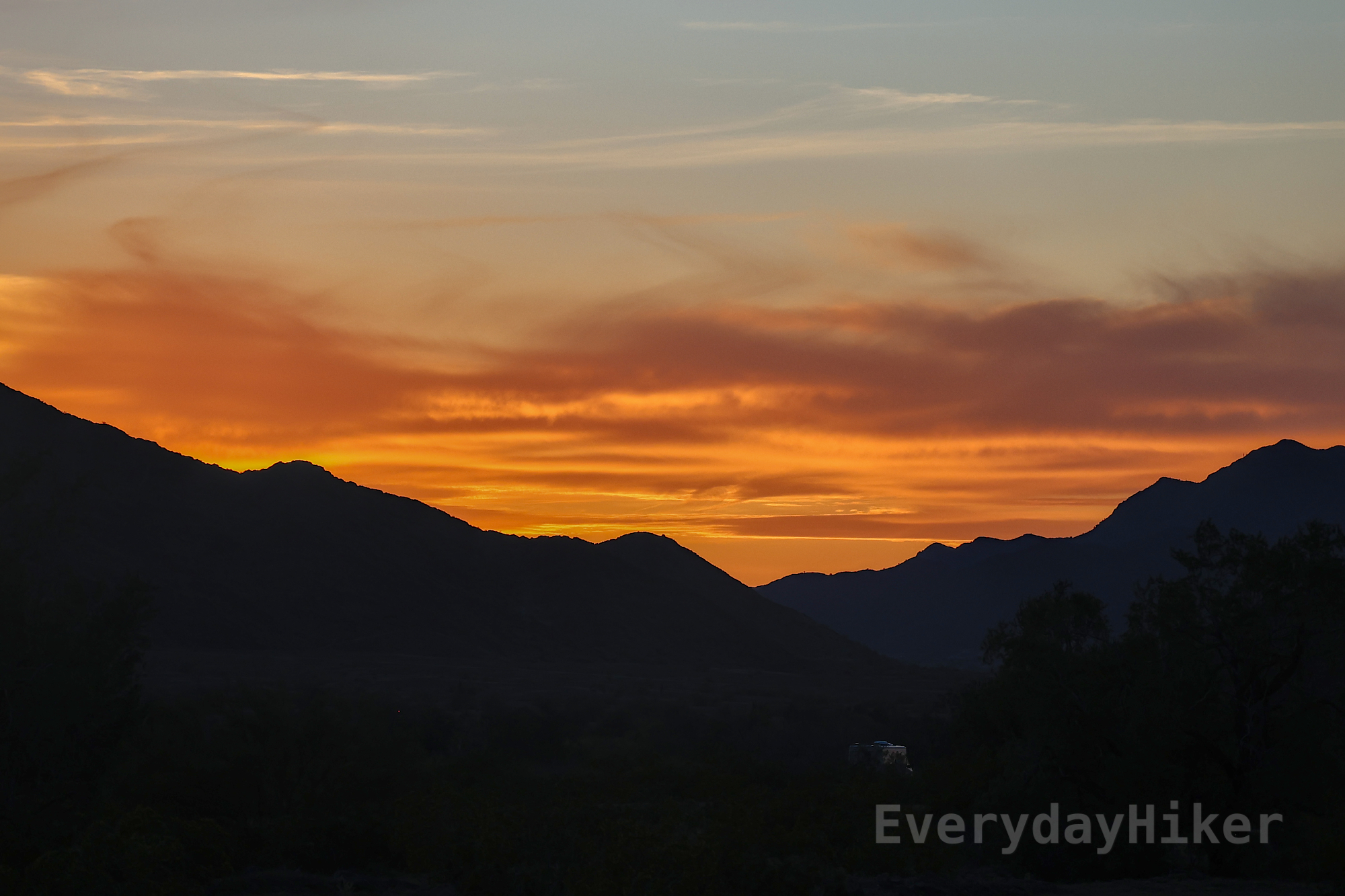 Sunset taken from the Ironwood National Monument BLM land near the Saguaro National Park. Two shadowed mountain ridges may be seen leading from opposite sides of the frame in the distance, while a brilliant orange glow burns above them. Small bands of clouds may be seen in the sky which turns to blue along the upper frame.