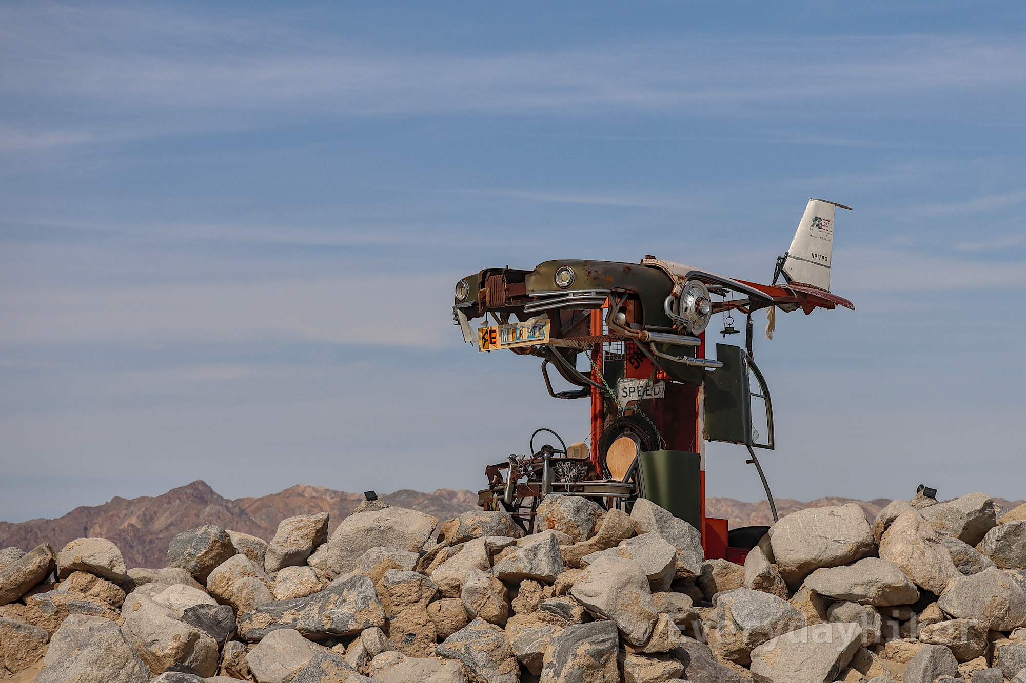 An interesting sculpture of a 50s style car converted into a small floating plane. It is surrounded by small boulders and in the distance a mountain range may be seen. License plates cut and pieced together form the word 'REINCARNATION.'
