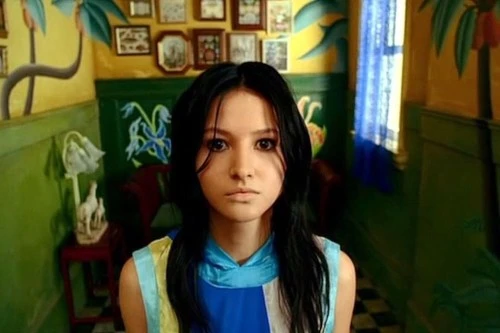 A screencap of Mimi from the movie survive style 5+. It shows a young woman with long black hair, looking somewhat dead-eyed into the camera. She's wearing a very colourful top, somewhat in the style of David Hockney. The room inteh background is similarly styled. The walls painte dark green for the lower half, and yellow for the top part, with palms painted on, a window with blue see-through curtains. And some pictures hanged in the background.